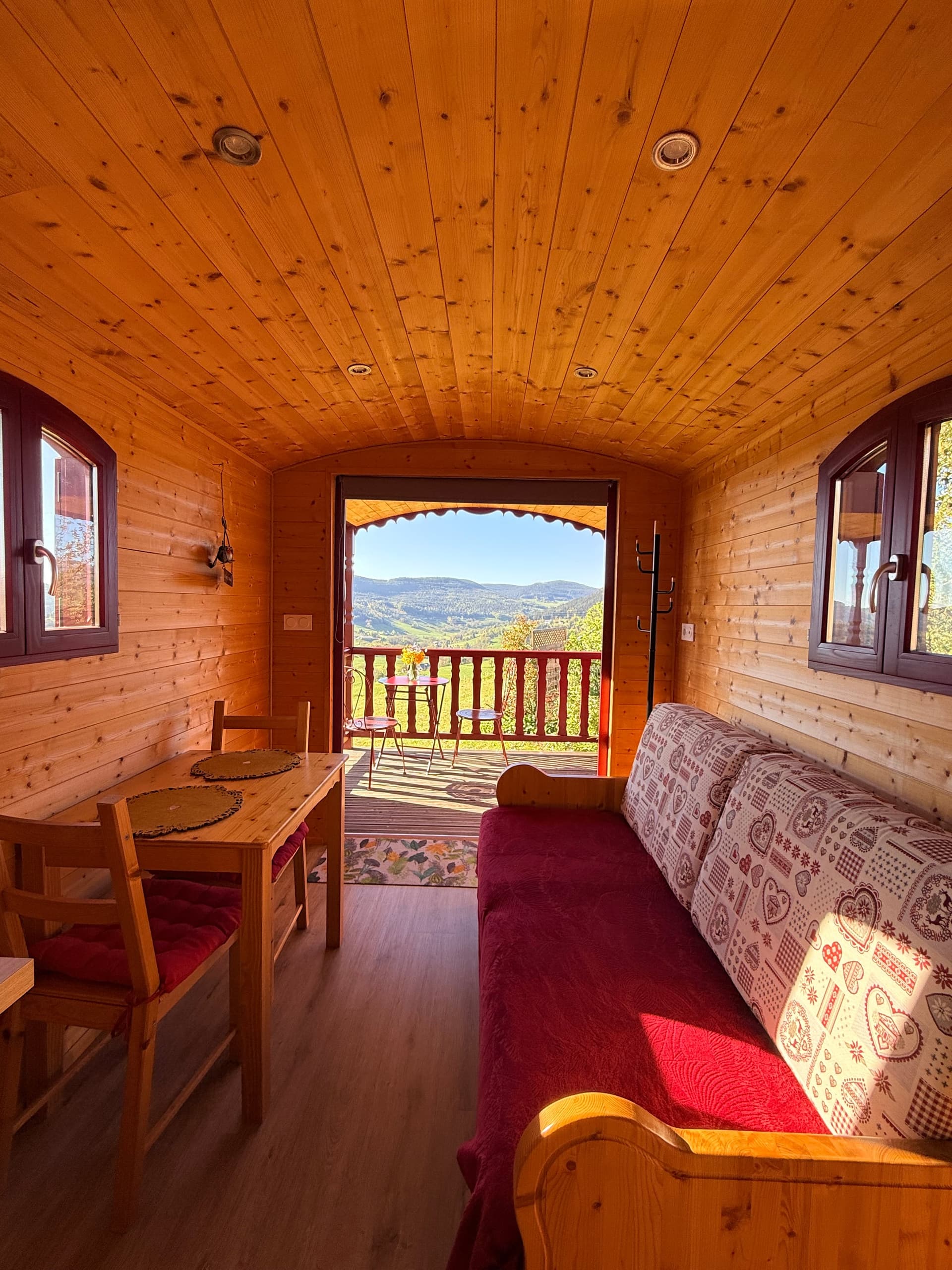 Séjour de la roulotte : banquette, table et porte ouverte sur la terrasse et la vallée.